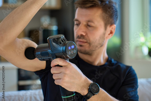 Athletic man using percussion massage device indoors. Man applies massage gun to arm muscle while sitting in living room, recovery therapy after exercise. Concept of home fitness and relaxation