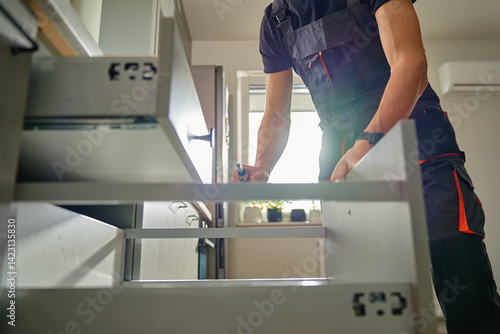 Man assembling kitchen drawer in modern cabinet using screwdriver. Handyman installing and adjusting drawer slide mechanism in contemporary kitchen. Furniture installation