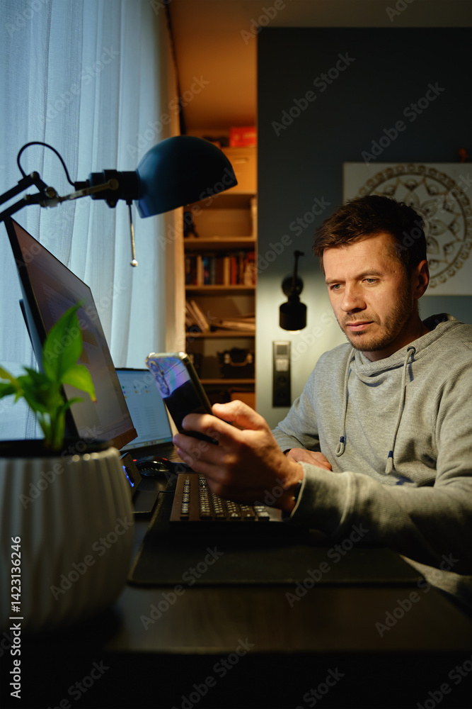Tired freelancer procrastinating at home office, scrolling phone instead of working. Unproductive man using smartphone at home desk. Concept of procrastination and distraction in remote work