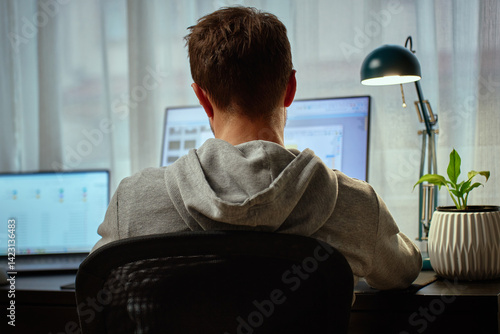 Obraz na płótnie Man in gray hoodie working at computer desk with dual monitors and lamp in background. Freelancer working remotely at home office. Concept of remote work and productivity w salonie