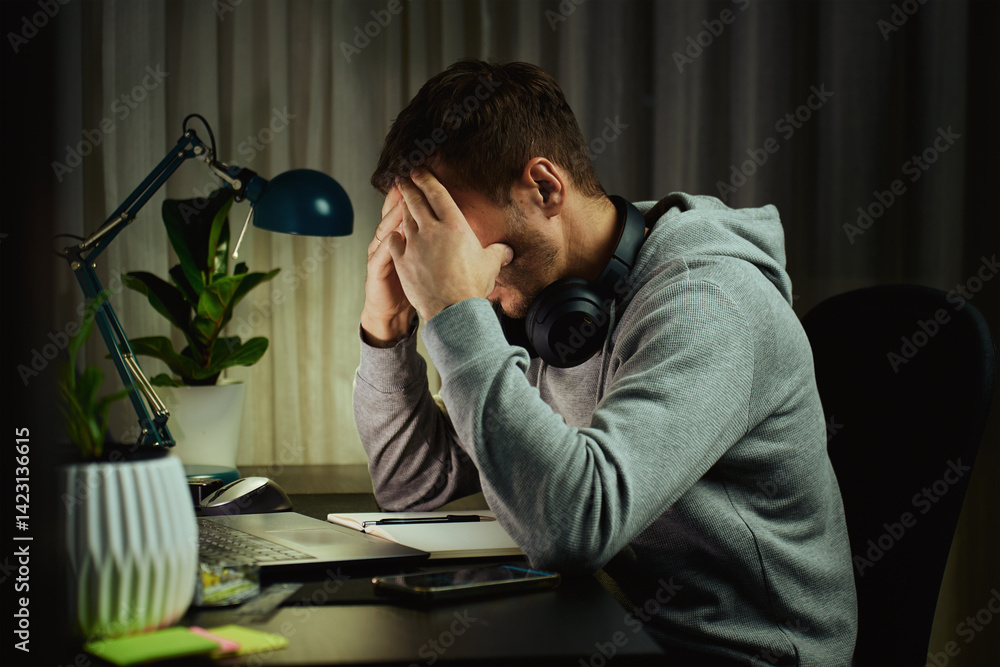 Tired man rubs eyes while working at computer. Fatigued freelancer working late at home office. Concept of stress and mental fatigue during remote work