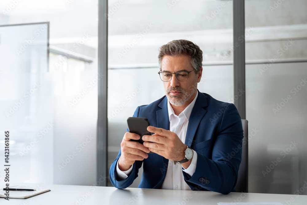 Focused Indian mature businessman in suit and eyeglasses holding smartphone sitting in office, working on device. Middle eastern arab or latin senior business manager man ceo using cell phone mobile Focused Indian mature businessman in suit and eyeglasses holding smartphone sitting in office, working on device. Middle eastern arab or latin senior business manager man ceo using cell phone mobile