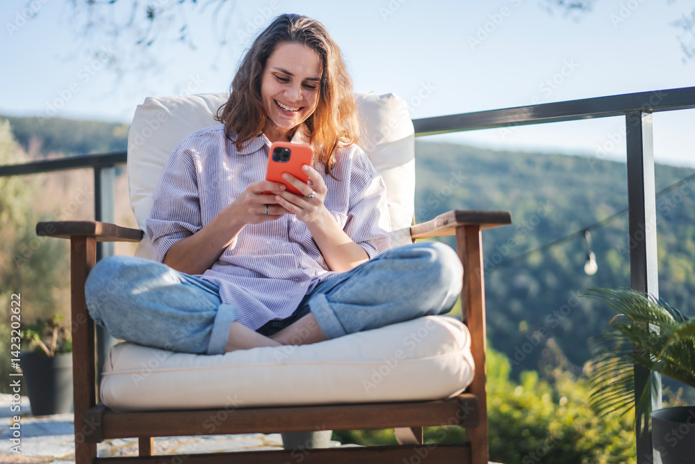 Happy young woman using mobile phone while sitting on the patio in summer.