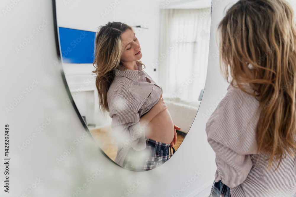 Young attractive pregnant woman looking at herself in the mirror while spending time at home