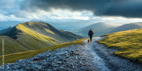 A lone hiker walks a mountain trail with their dog, enjoying a scenic landscape under a dramatic sky. Perfect for travel and adventure brochures.