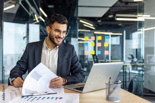 A smiling businessman reviews documents, with a laptop open on his desk in a modern office setting, ready to work.