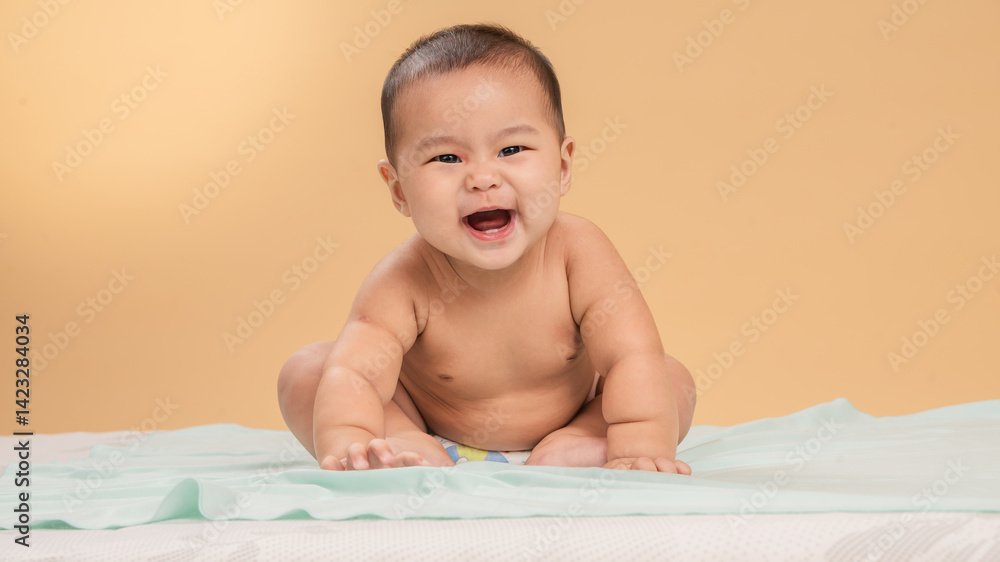 Adorable Asian baby wearing only a diaper, sitting and smiling brightly on a soft mint-colored sheet with a beige studio background, showing joyful and playful expression