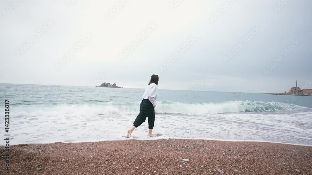 A cinematic handheld shot of a middle-aged woman walking along the beach barefoot on a cloudy day A cinematic handheld shot of a middle-aged woman walking along the beach barefoot on a cloudy day