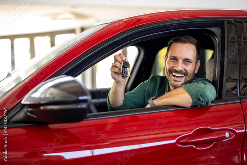 Handsome man raising hand up with the car key and smiling at camera, showing key from his brand new car, copy space. Man buying auto at luxury dealership salon