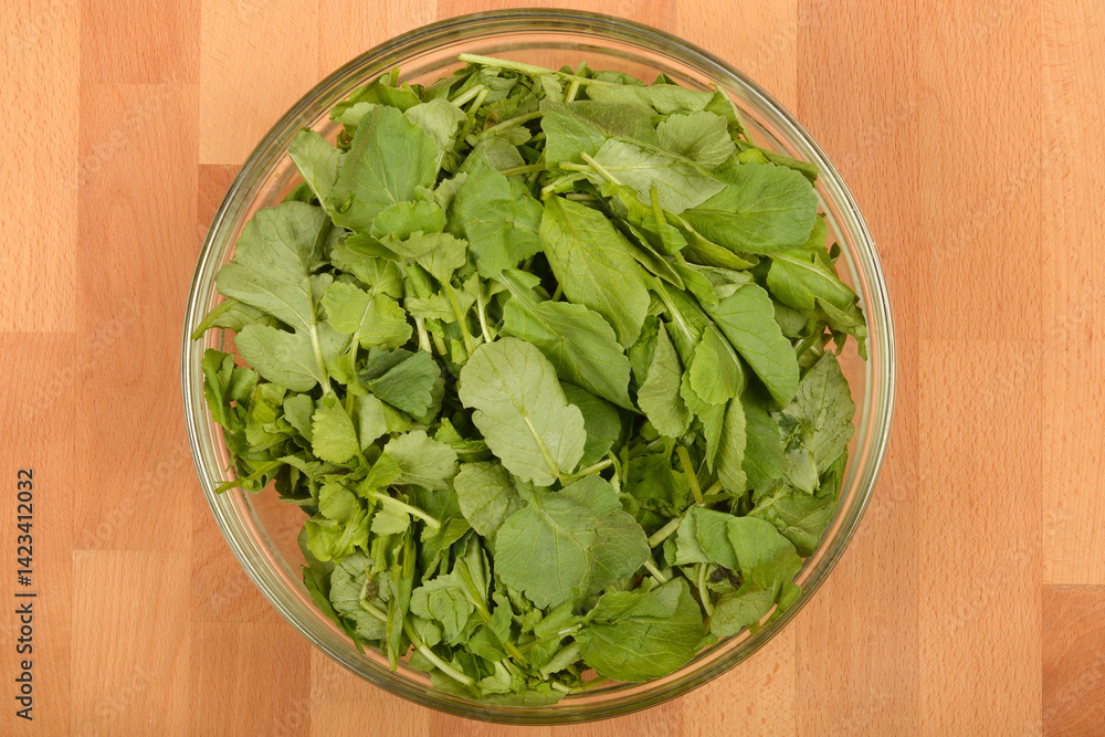 A clear glass bowl filled with fresh, vibrant green radish leaves sits on a light wooden surface, captured from a top-down perspective