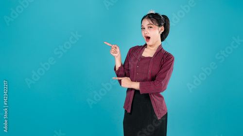 Tableau sur toile Young woman in traditional Javanese lurik costume smiling confidently while pointing sideways with both hands, conveying a cheerful and persuasive expression in studio setting