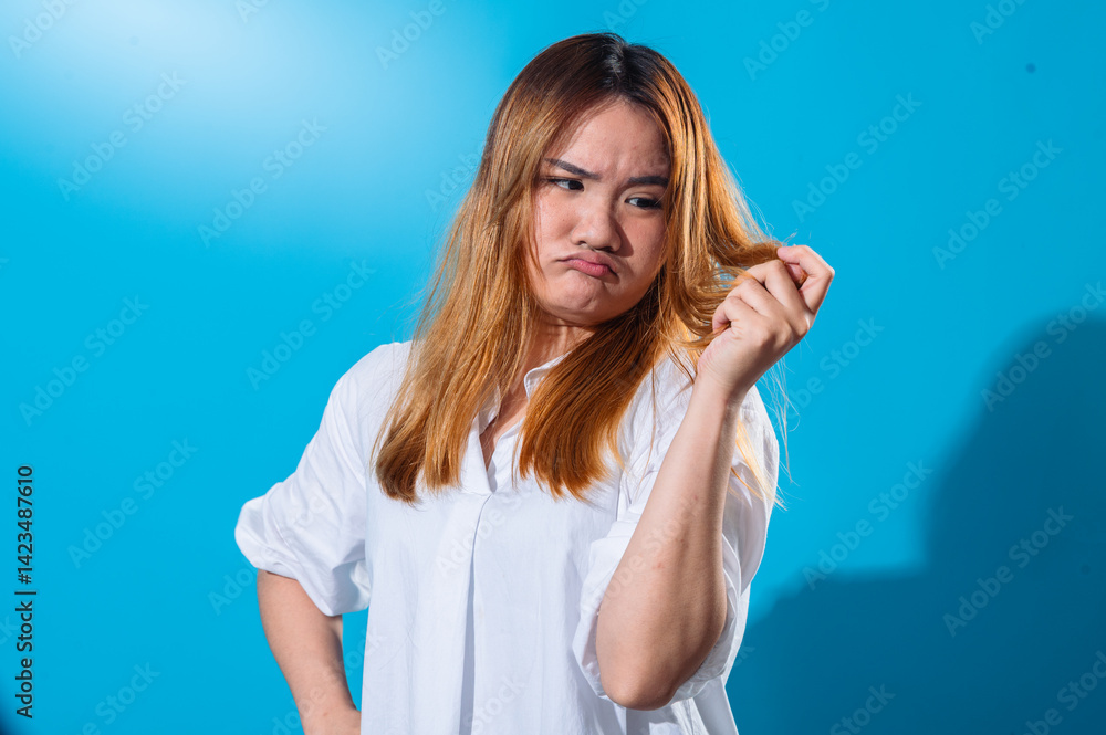 Fototapeta premium Asian woman in a white shirt looking at her hair with a concerned and displeased expression, examining split ends or dryness, standing against a solid blue background in studio light