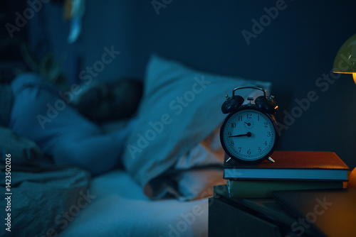 Person peacefully sleeping in bed with alarm clock and lamp on bedside table. Soft dim light creating calm and restful atmosphere
