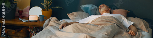фотографія Elderly man resting in a cozy bedroom setting with bedside lamp and plants, creating a serene and comforting atmosphere