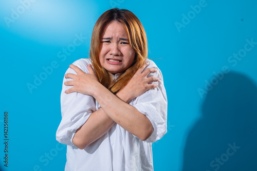 Asian woman in a white shirt hugging herself tightly with a fearful and anxious expression, standing against a bright blue background, conveying a sense of cold, fear, or emotional discomfort