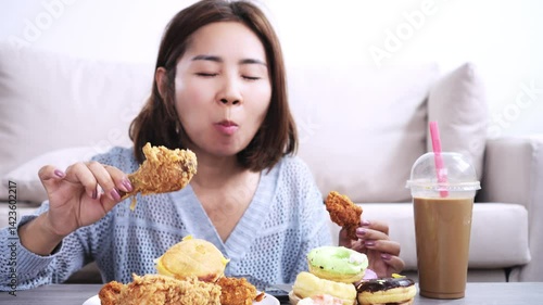 asian woman happily eating fast food like fried chicken, with donuts, cake, burgers, and sweet coffee on the table, showing unhealthy eating habits with oily, salty, and sugary foods and drinks