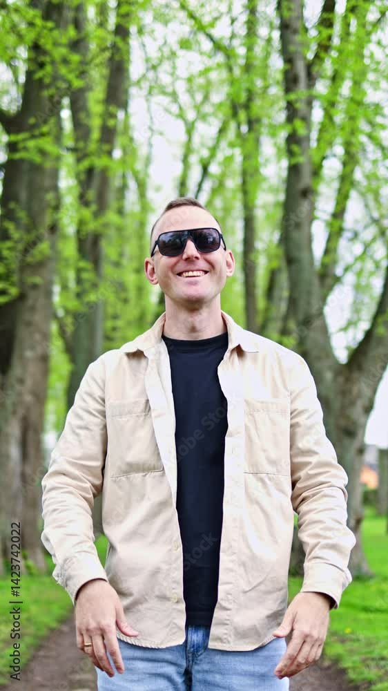A cute man in sunglasses and a beige jacket is walking in the park. A cheerful man looks into the camera and calls to follow him. The guy is walking in the spring park. A cute man in sunglasses and a beige jacket is walking in the park. A cheerful man looks into the camera and calls to follow him. The guy is walking in the spring park.