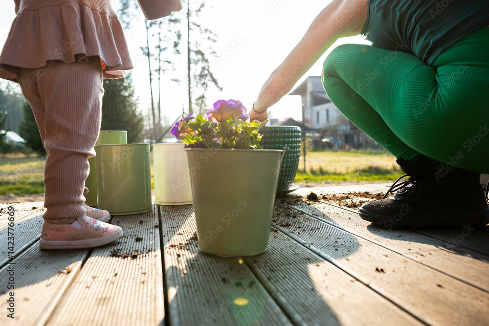An adult in green leggings and a child in pink clothing plant flowers on a wooden deck. Pots and soil are scattered, with trees and a building in the background.