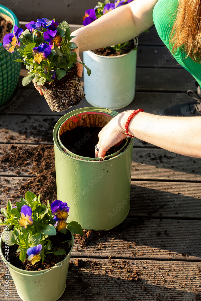 A person in a green shirt plants flowers in a light green pot on a wooden deck, with soil, vibrant purple and yellow flowers, and sunlight present.