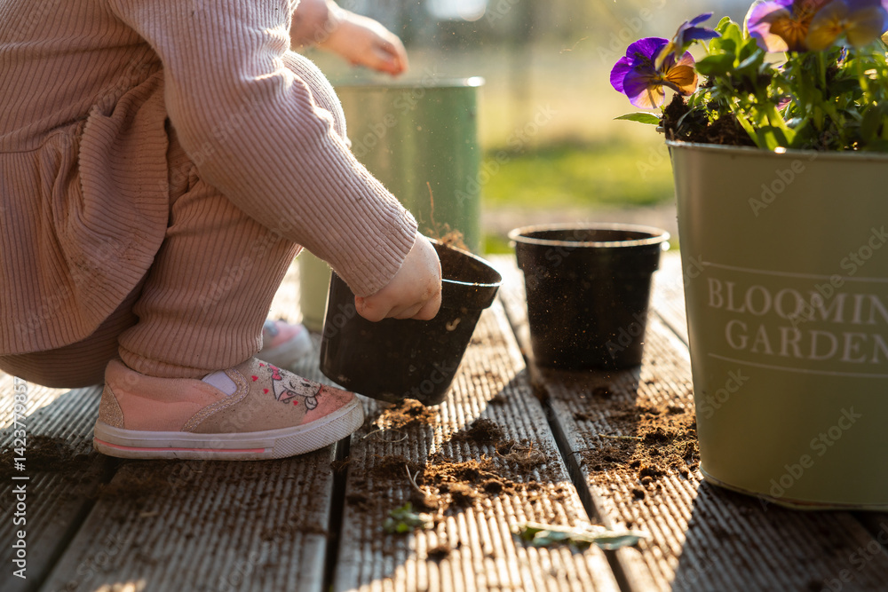 A child in pink shoes and light clothing works with soil in black pots on a wooden deck. A green container with purple and yellow flowers is nearby.