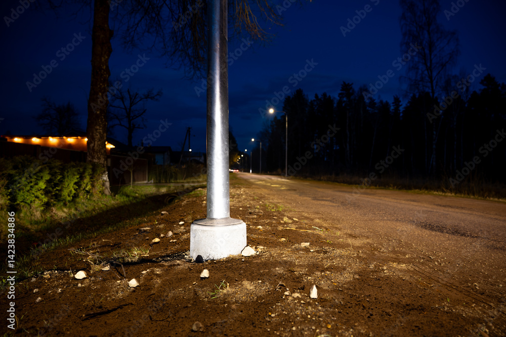 A rural road at night features a metal streetlight pole in the foreground, sparse trees, a fence, and a faint glow from a building with lit windows.