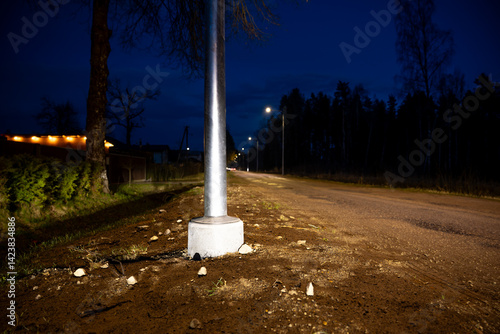 Obraz na płótnie A rural road at night features a metal streetlight pole in the foreground, sparse trees, a fence, and a faint glow from a building with lit windows. w salonie