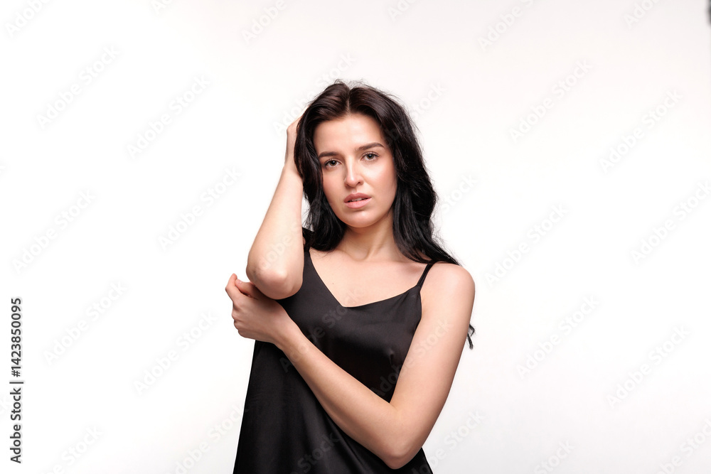 Young woman in black dress poses with hand on arm against plain background in bright lighting during photo shoot Young woman in black dress poses with hand on arm against plain background in bright lighting during photo shoot