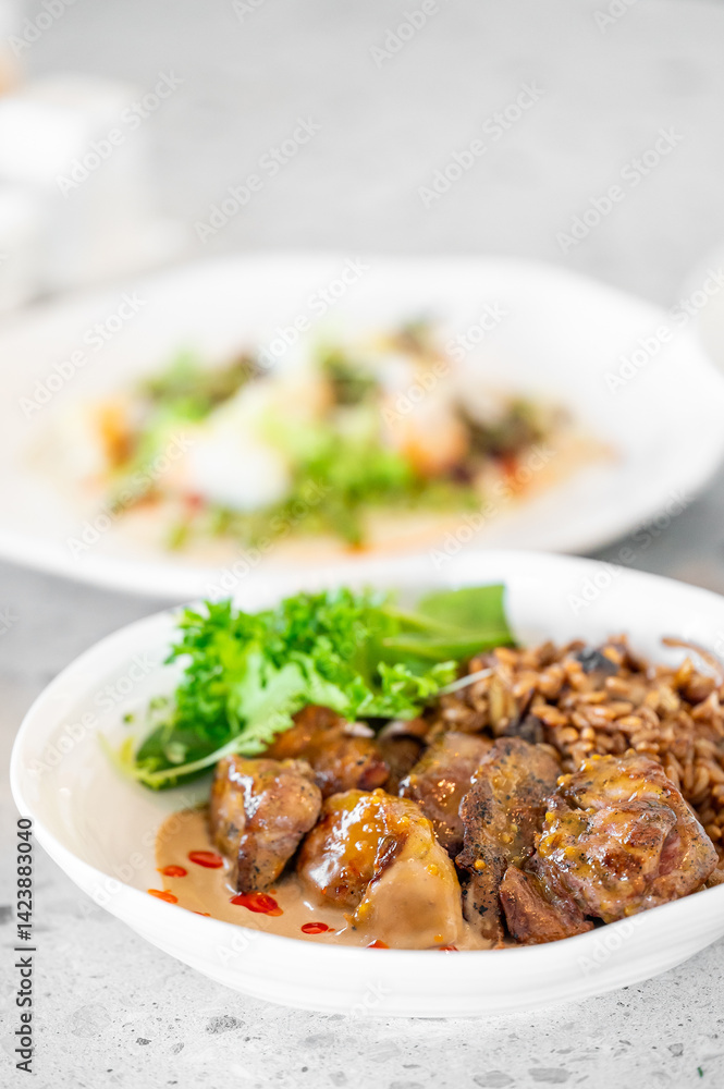 A vibrant dish featuring succulent meat pieces, grains, and fresh greens, elegantly presented in a white bowl. A blurred background displays another beautifully plated meal, enhancing the dining A vibrant dish featuring succulent meat pieces, grains, and fresh greens, elegantly presented in a white bowl. A blurred background displays another beautifully plated meal, enhancing the dining