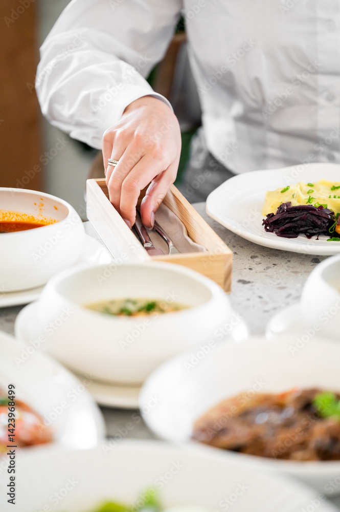A close-up of a person's hand reaching for utensils from a wooden holder, surrounded by a variety of delicious dishes, creating an inviting dining atmosphere. A close-up of a person's hand reaching for utensils from a wooden holder, surrounded by a variety of delicious dishes, creating an inviting dining atmosphere.