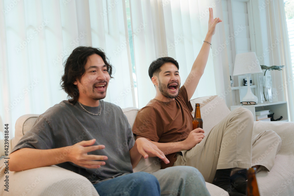 Fototapeta premium Two Asian Male friends are having a fun game night in a living room, sharing drinks and cheering excitedly as they play together.