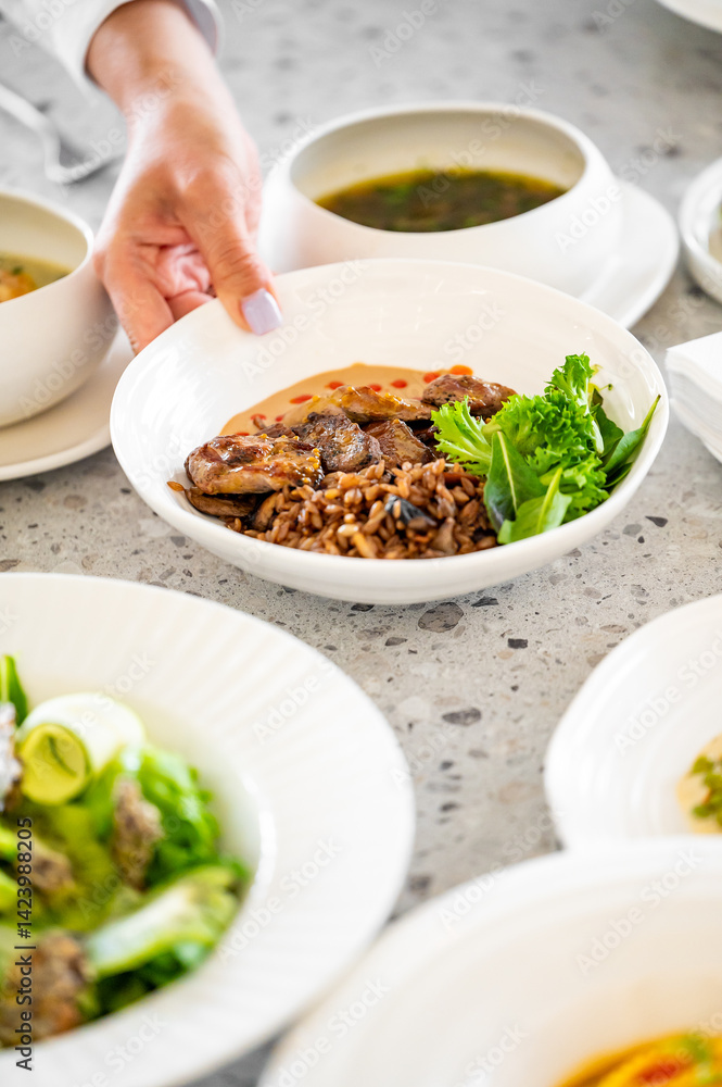 A white bowl with cooked meat, rice, and lettuce placed on a speckled countertop, surrounded by dishes of salads and soup, showcasing a healthy and appetizing meal arrangement. A white bowl with cooked meat, rice, and lettuce placed on a speckled countertop, surrounded by dishes of salads and soup, showcasing a healthy and appetizing meal arrangement.