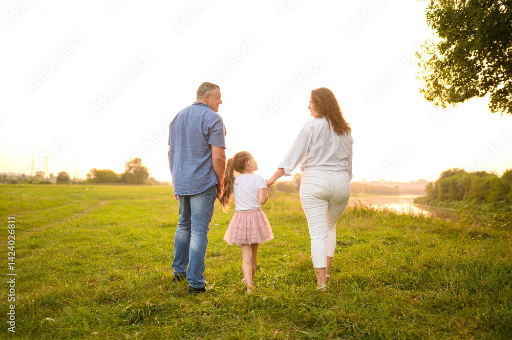 Back view of family walking together in summer park at sunset. Mom, dad, daughter walking in meadow holding hands, smiling happily looking at each other. Family, love and parenthood concept. Back view of family walking together in summer park at sunset. Mom, dad, daughter walking in meadow holding hands, smiling happily looking at each other. Family, love and parenthood concept.