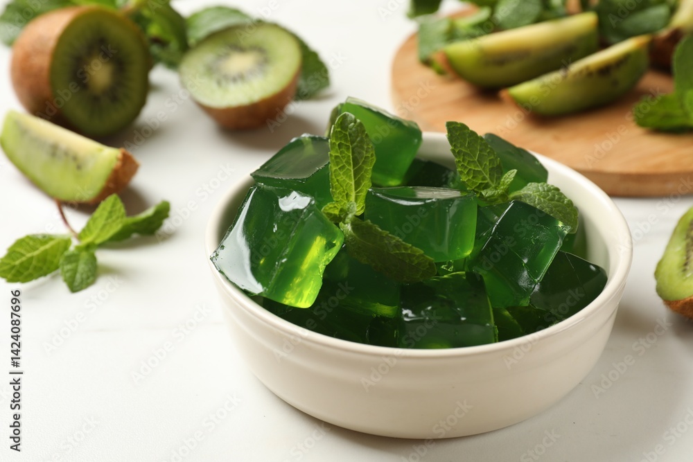 Obraz premium Delicious green jelly cubes in bowl, mint and kiwi on white table, closeup