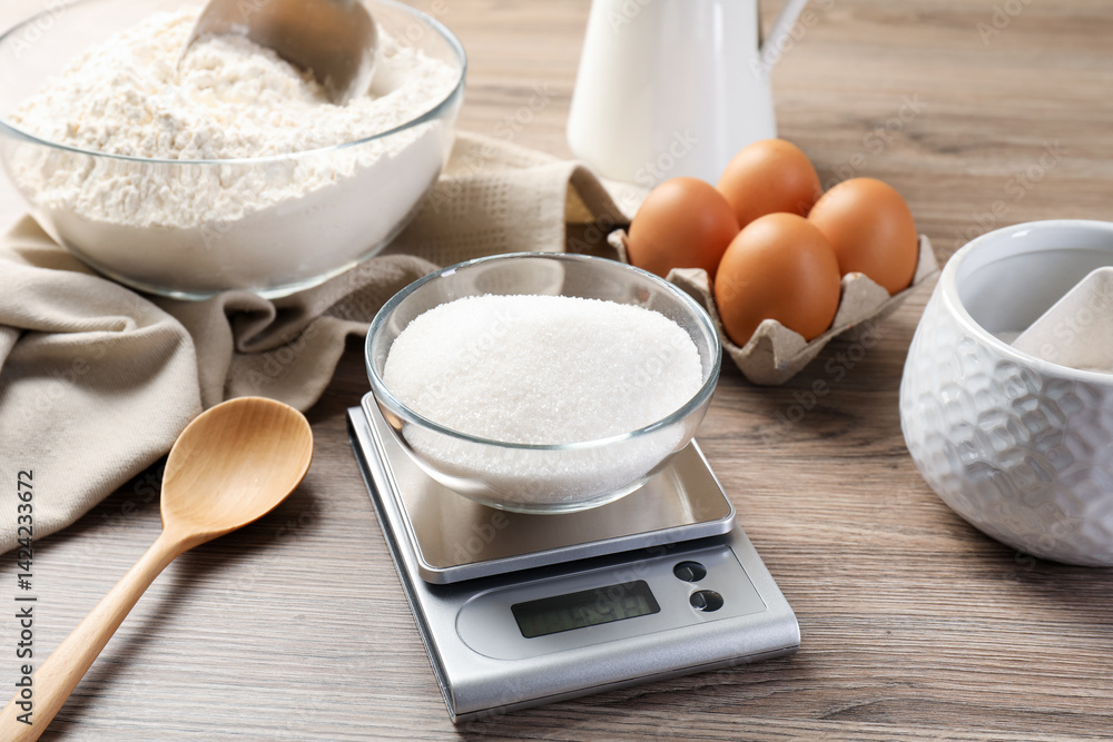 Digital kitchen scale with bowl of sugar among other products on wooden table