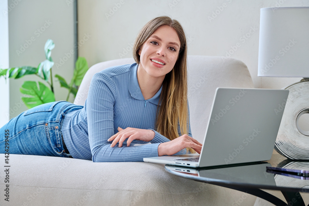 Portrait of young woman lying on sofa at home with laptop looking at camera