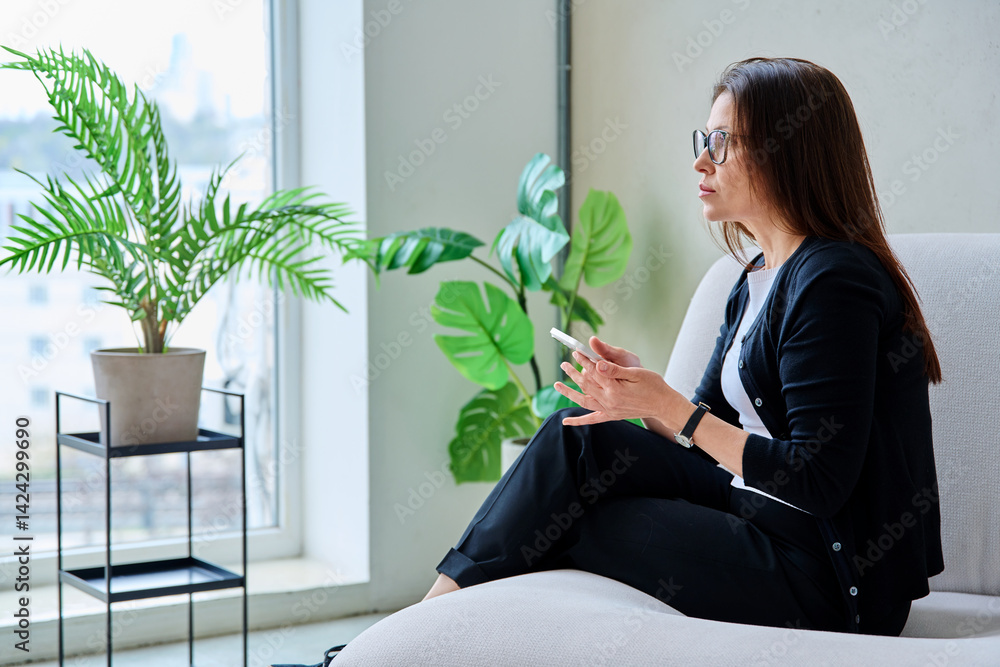 Serious anxious middle aged woman looking out window sitting on sofa