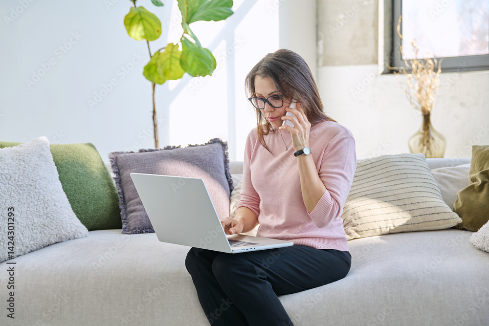 Middle aged woman sitting on sofa with laptop smartphone, in living room interior. Mature female using