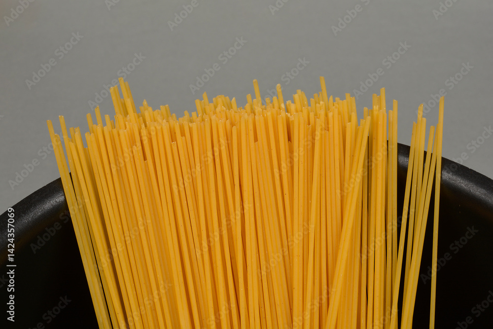 A bundle of dry, uncooked spaghetti stands upright in a black pot against a neutral background