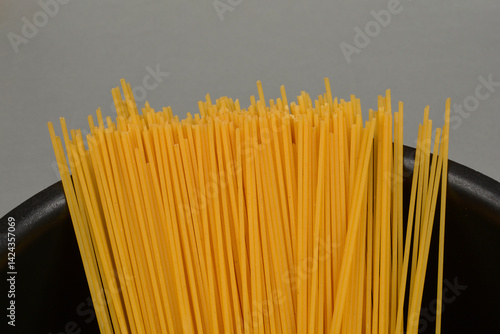 A bundle of dry, uncooked spaghetti stands upright in a black pot against a neutral background