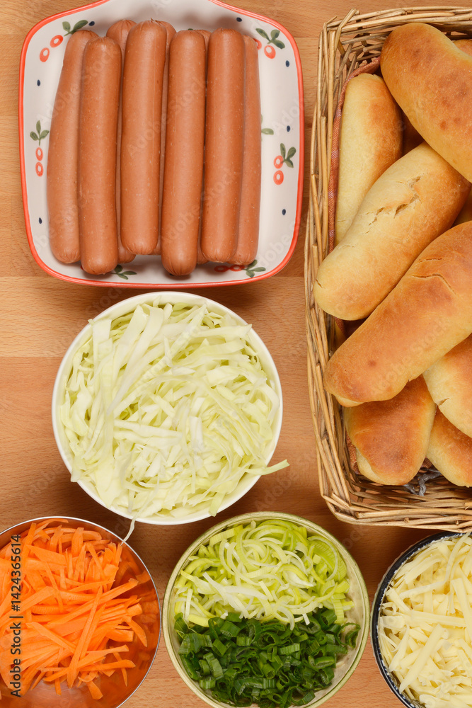 An overhead shot showcases the components for preparing hot dogs: buns in a basket, frankfurters on a serving dish, shredded cabbage and cheese, and chopped leeks with scallions