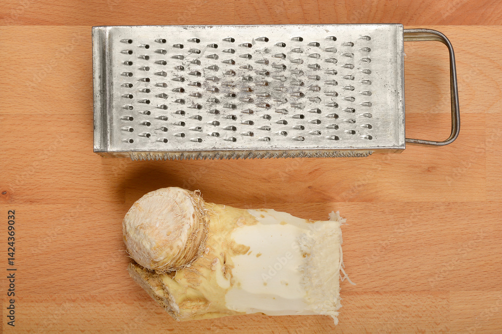 A piece of horseradish root is positioned below a meta grater on a light brown wooden board