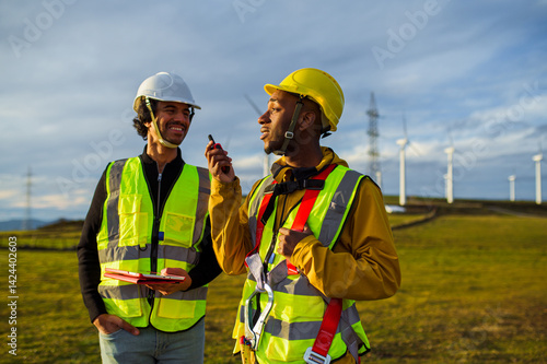 Two multi ethnic engineers communicating using a walkie talkie while working in a wind turbine field, wearing safety vests and helmets
