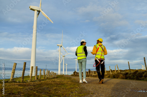 Obraz na płótnie Two engineers walking alongside wind turbines, engaged in discussion while wearing safety vests and helmets in a rural landscape w salonie