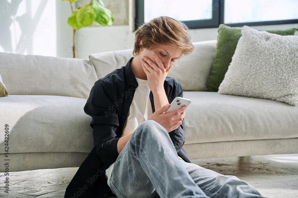 Upset sad teenager guy looking at smartphone screen sitting on floor at home