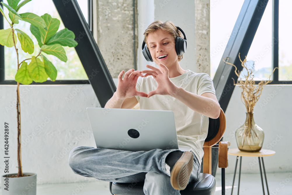 Happy teenage boy with headphones using laptop for video chat, making heart gesture