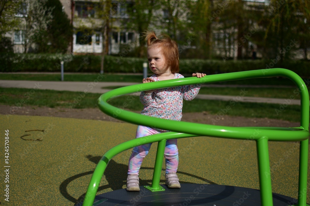 Fototapeta premium Baby girl standing on spinning equipment in children playground in city park in warm weather spring. Toddler girl having fun spinning around in public playground in sunny day spring.