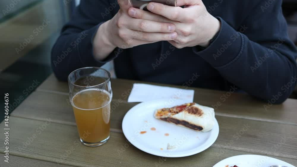 Person using smartphone while eating grilled burrito and drinking beer at a wooden table during a casual outdoor meal. Person using smartphone while eating grilled burrito and drinking beer at a wooden table during a casual outdoor meal.