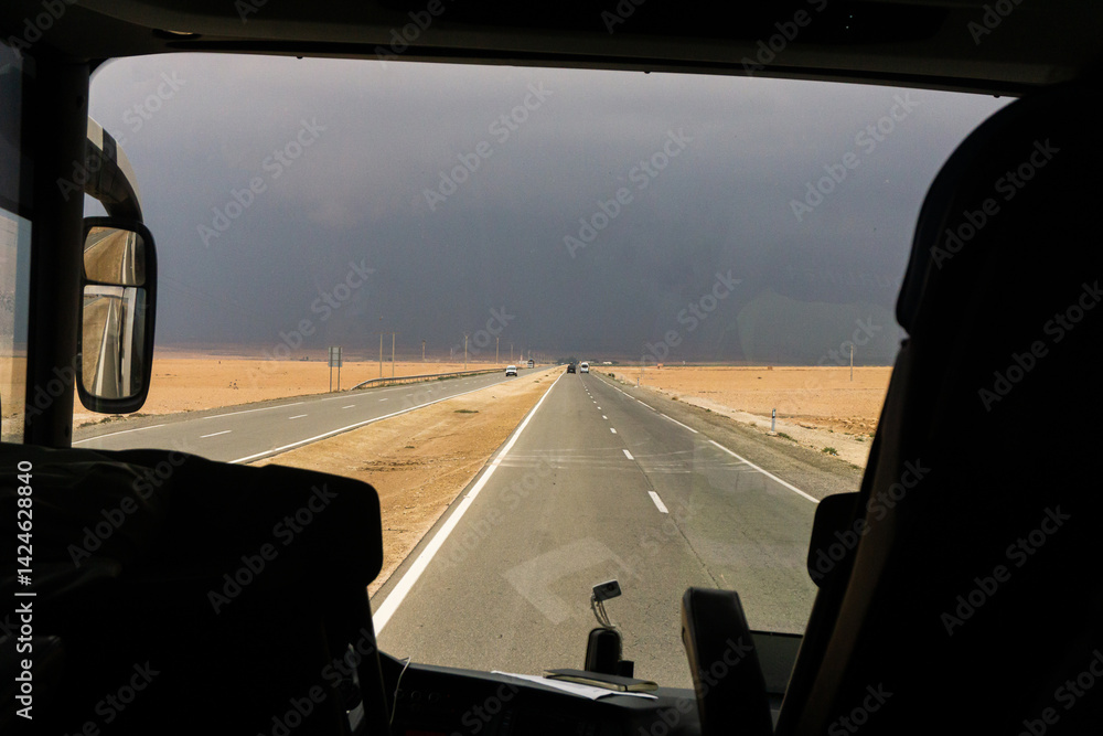Desert road view from bus. Stormy sky horizon. Travel in Morocco. Long straight highway. Empty sandy landscape. Dramatic road trip. Adventure drive. Transport journey. Asphalt desert route.