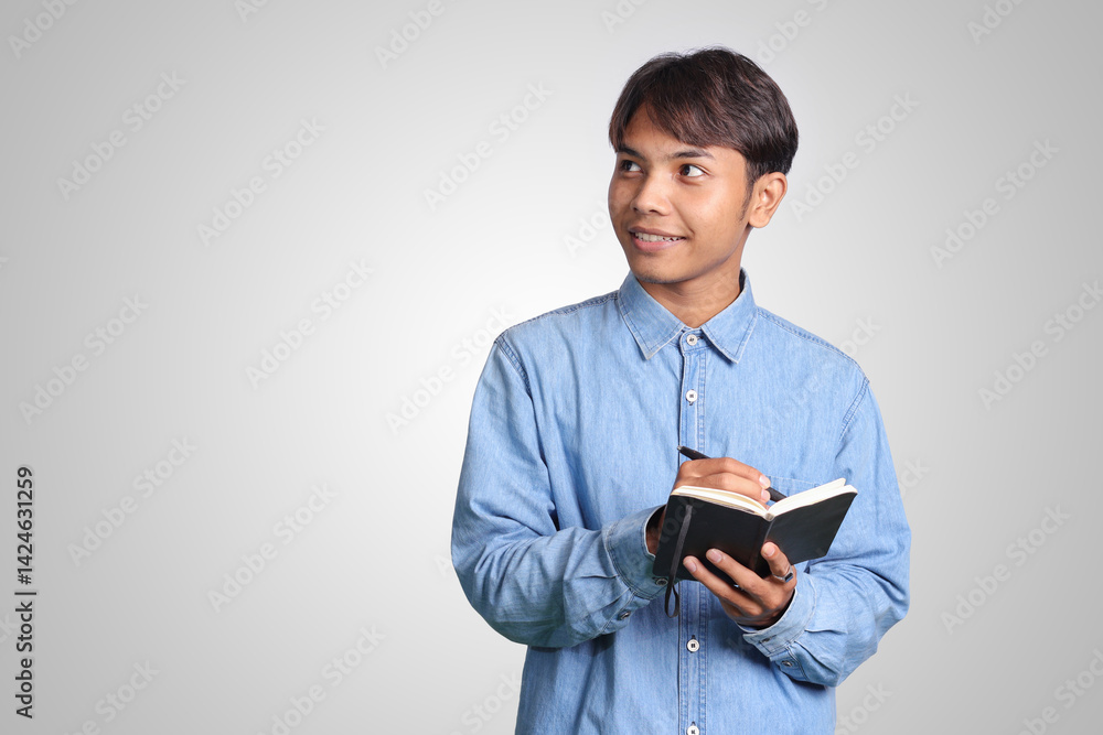 Portrait of confused Asian man in blue shirt writing on his notes while thinking about idea. Isolated images on white background Portrait of confused Asian man in blue shirt writing on his notes while thinking about idea. Isolated images on white background