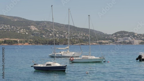 Several sailboats anchored in calm blue sea with mountains and coastal town in background. Sunny summer day in Mallorca, peaceful maritime scene, themes of travel, vacation, sailing, and nature.